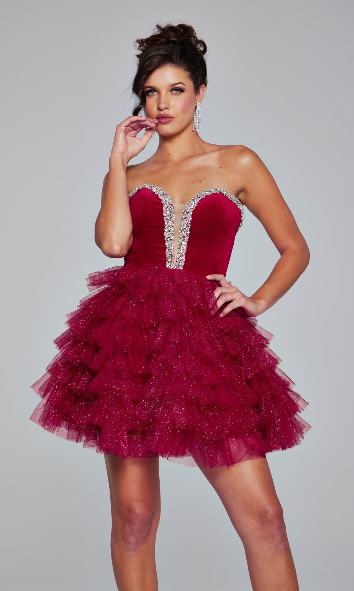 A woman wears the Jovani 40352 Strapless Short Glitter Hoco Dress in deep red, featuring a ruffled tulle skirt and rhinestone-embellished bodice, standing against a plain gray background.